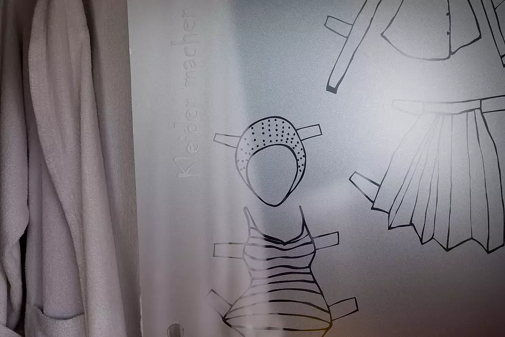 Detail. Auf einer Schrankschiebetür befindet sich eine Grafik von Kleidungstücken: Badeanzug, Badekappe, Faltenrock. Im linken Teil des Bildes sieht man Details von echte Kleidungsstücken aus dem Schrank.