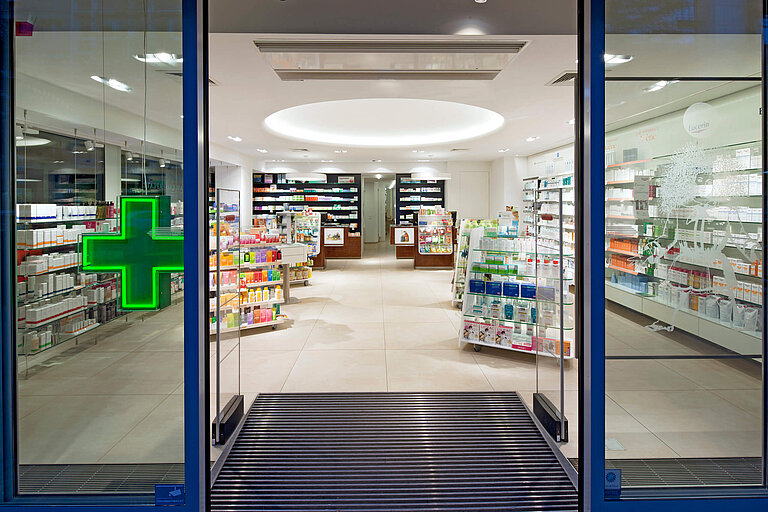 Außenansicht durch Fensterfront. Warensicherungskontrolle und leuchtendes Apothekenkreuz.