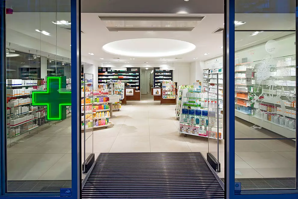 Außenansicht durch Fensterfront. Warensicherungskontrolle und leuchtendes Apothekenkreuz.